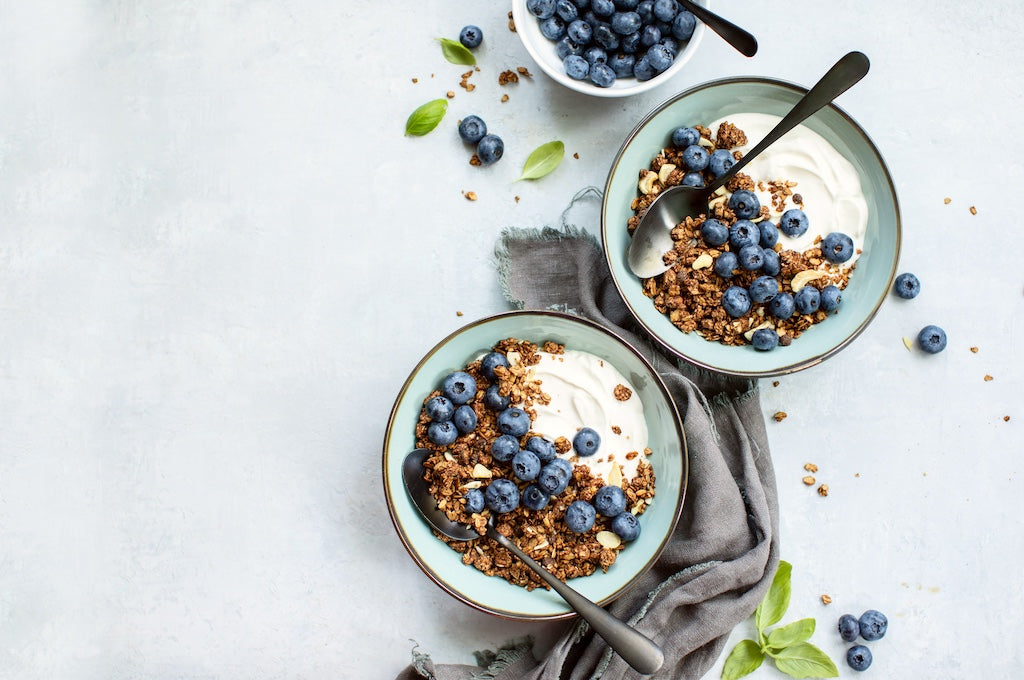 Two bowls of yogurt with granola and fresh blueberries, each with a spoon. Also visible on the table is a small bowl of blueberries and scattered leaves.