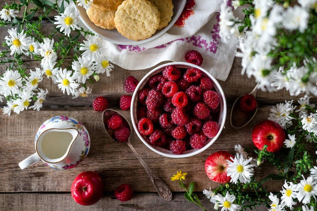 A bowl of fresh raspberries sits on a wooden table, surrounded by apples, cookies, a jug of cream, and white flowers.