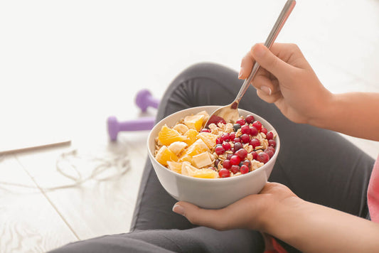 Eine Person hält eine Schüssel Haferflocken mit Orangenscheiben, Banane und roten Beeren – und genießt dieses wärmende Frühstück, während sie auf dem Boden neben lila Hanteln sitzt, perfekt für alle, die abnehmen möchten.