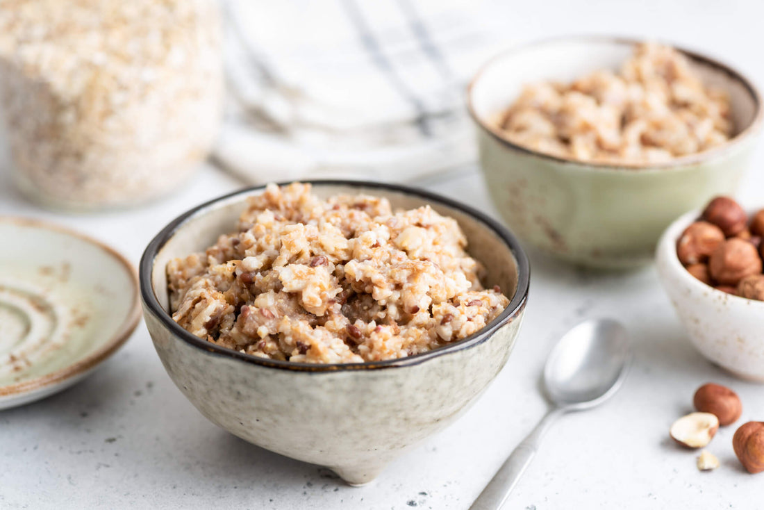 On a table there is a ceramic bowl filled with porridge next to a spoon, another bowl of porridge, a plate and a small bowl of hazelnuts.