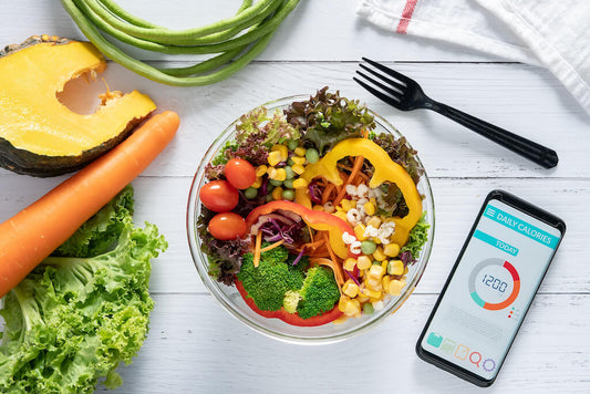 Eine Schüssel mit gemischtem Salat und Gemüse neben einem Smartphone mit einem täglichen Kalorienzähler, platziert auf einem weißen Holztisch mit frischem Obst und Gemüse drumherum.