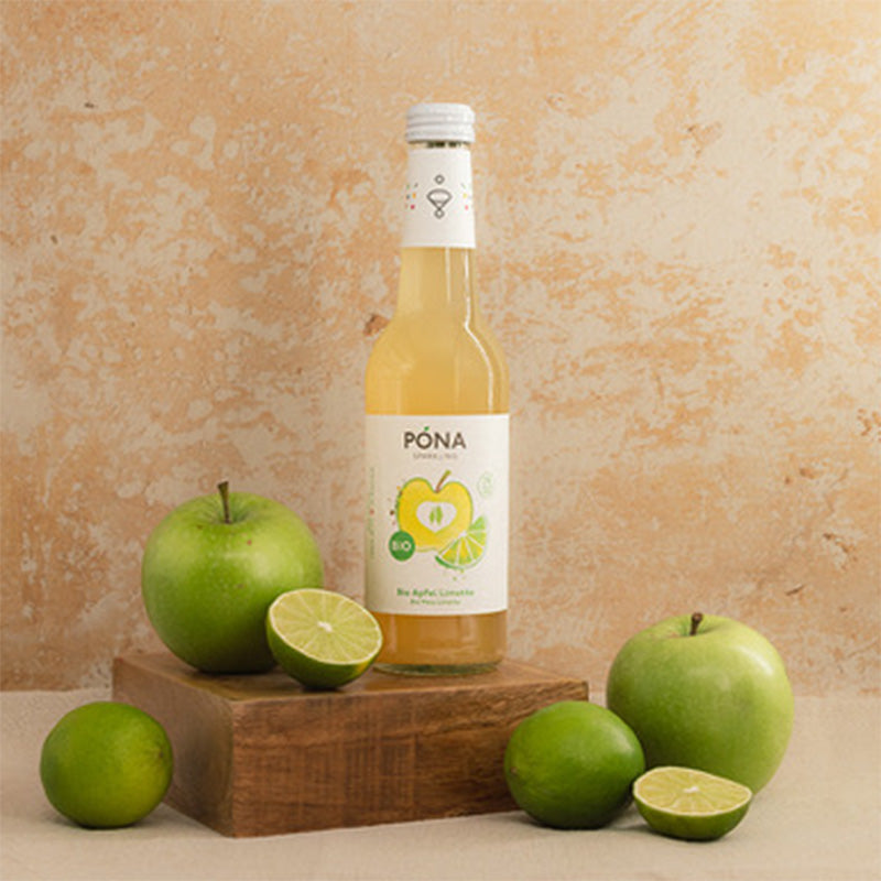 A bottle of PONA apple-lime drink on a wooden block, surrounded by green apples and lime slices, against a textured beige background.