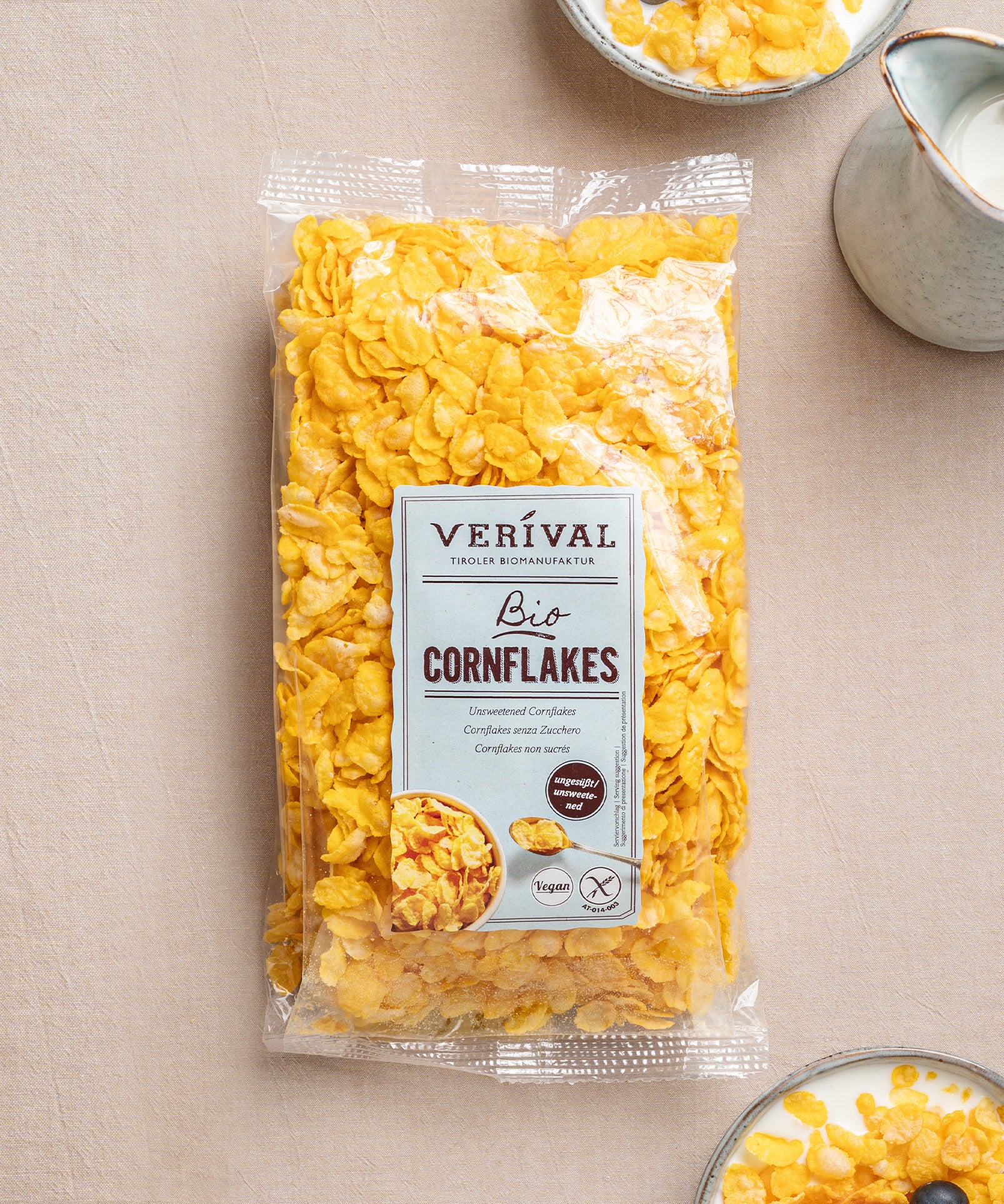 Eine durchsichtige Verpackung ungesüßter Cornflakes von Verival Bio liegt auf einer beigen Fläche, daneben eine Schüssel knusprige Cornflakes und ein Krug Milch.