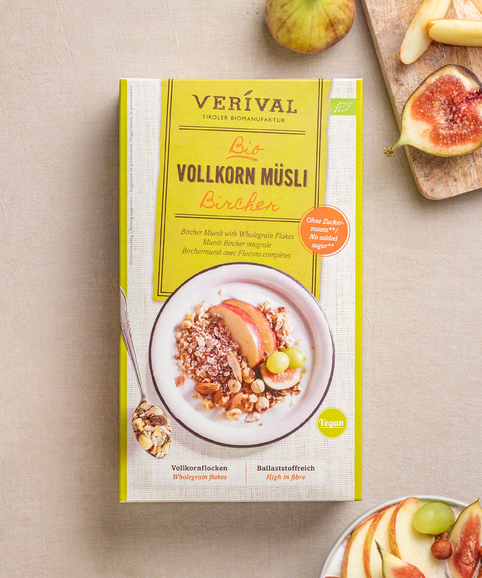Das vegane Bircher Vollkorn Müslis von Verival Bio wird mit Apfelscheiben, Nüssen, mit verstreuten Feigen und einem Löffel auf beigem Hintergrund gezeigt. Es ist reich an Ballaststoffen für einen gesunden Start in den Tag.
