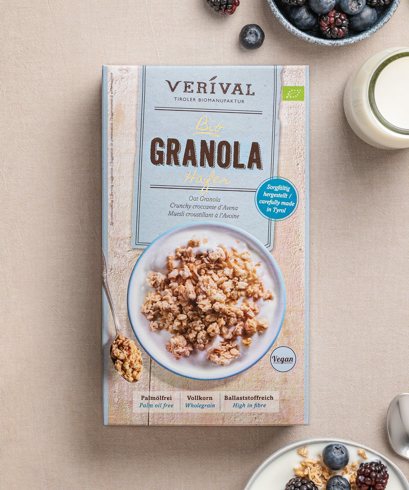 Auf einem Tisch steht eine Packung Hafer-Granola der von Verival Bio, dazu eine Schüssel knuspriges Granola, frische Beeren, vegane Kekse und ein erfrischendes Glas Milch.