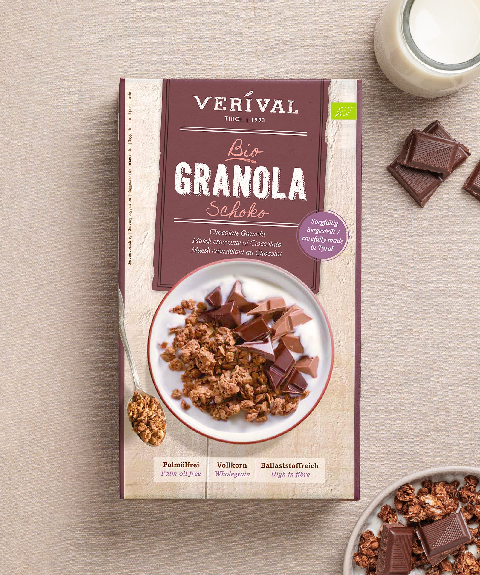 Auf dem Tisch steht eine Packung Schoko Granola der Verival Bio GmbH, dazu eine Schüssel knuspriges Schoko-Granola, ein Glas Milch und Schoko-Quadrate – ein ideales Frühstück voller Ballaststoffe.