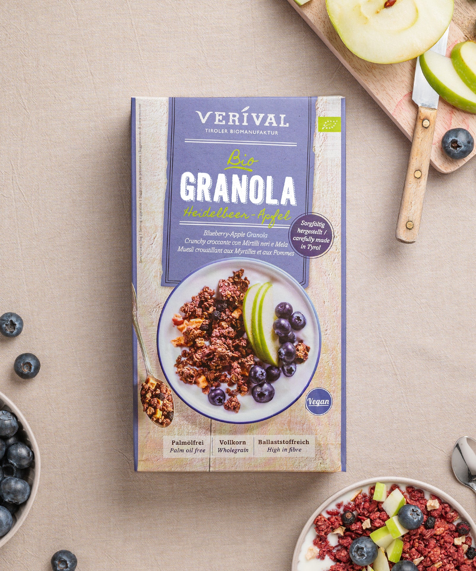 Eine Packung Heidelbeer-Apfel Granola von Verival Bio steht auf dem Tisch, umgeben von Schüsseln mit knusprigem Granola, Apfelscheiben und Heidelbeeren. Dieser köstliche vegane Leckerbissen ist ballaststoffreich und sorgt für einen gesunden Start in den Tag.