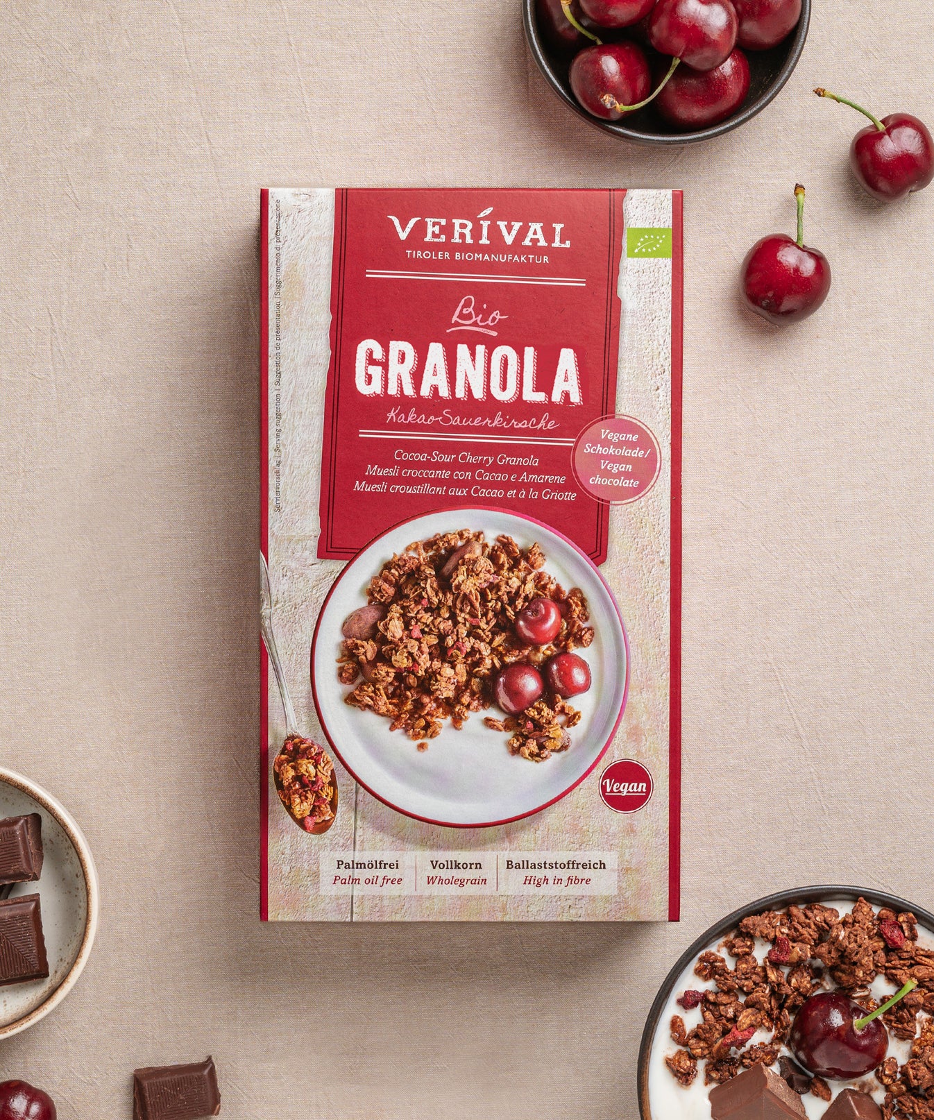 Eine Packung Kakao-Sauerkirsche-Granola mit Kirschen von Verival Bio, daneben Schalen mit veganem Granola, Kirschen und Schokostückchen auf einer hellen Tischdecke.