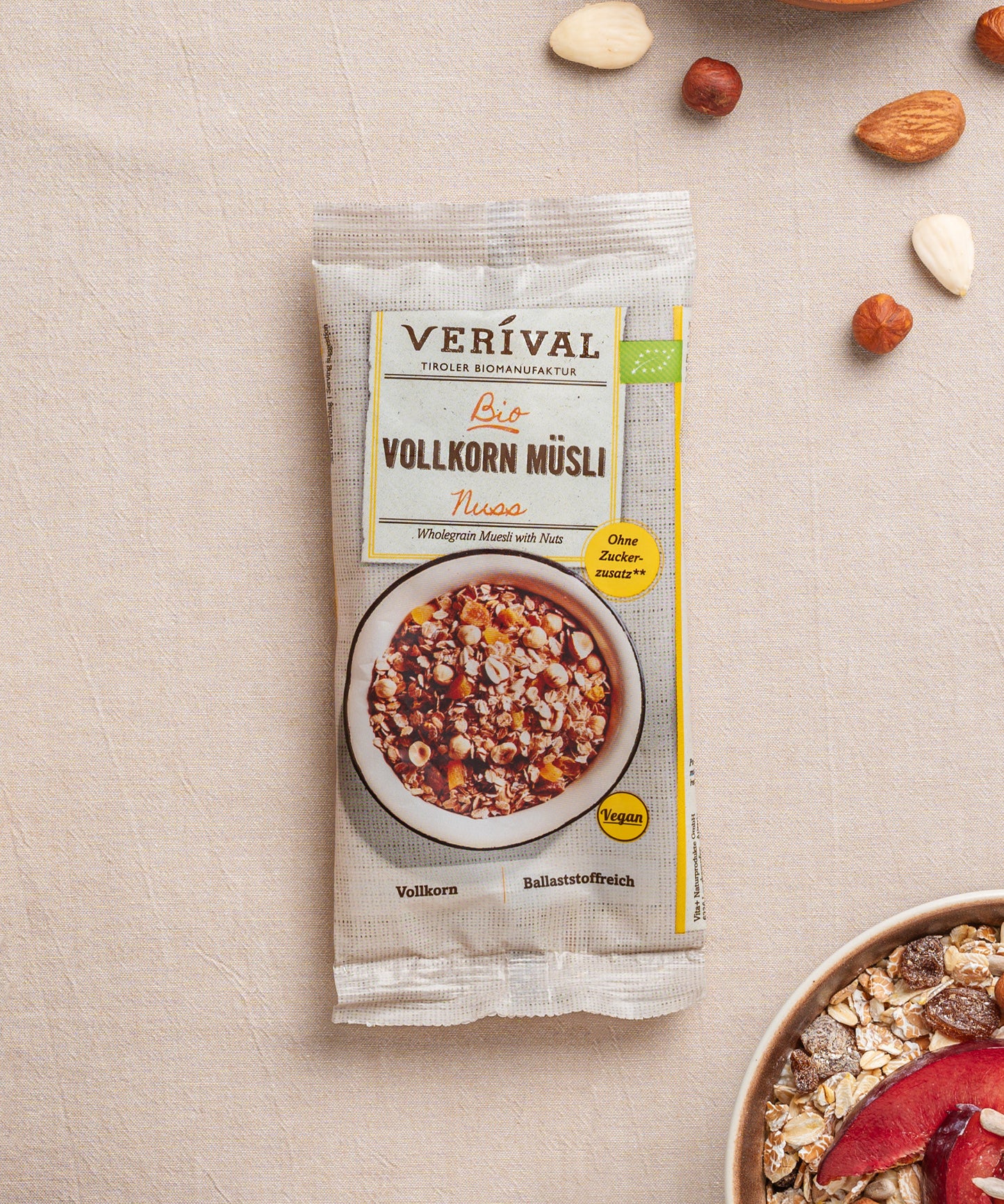Eine Packung Nuss-Vollkorn-Müsli der Verival Bio GmbH liegt auf einer strukturierten Oberfläche und unterstreicht so seinen veganen Reiz. Die Schachtel enthält eine Schale ballaststoffreiches Müsli mit Nüssen, dazu verstreute Nüsse und einen Teil einer weiteren Schale.