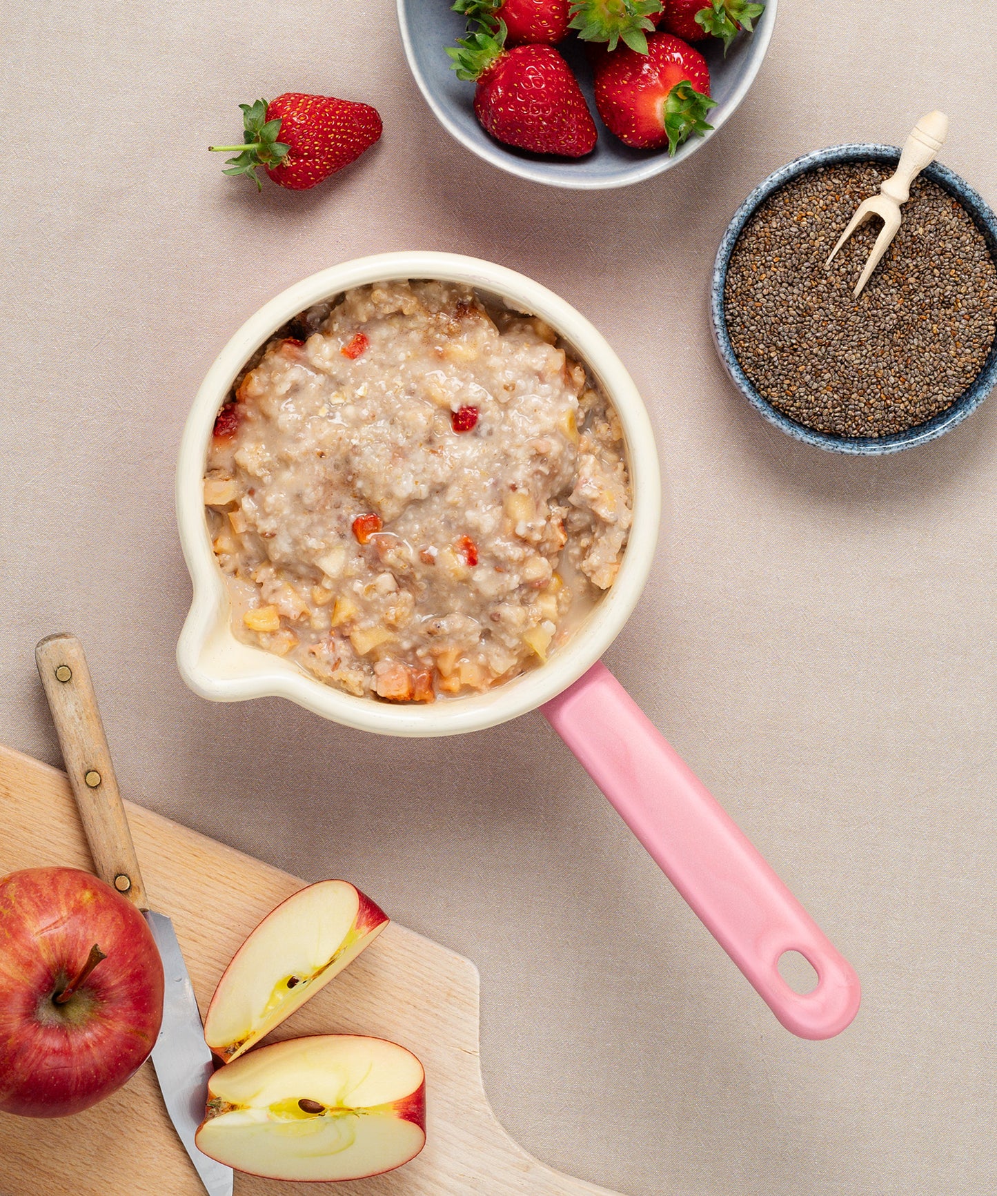 Der charmanter Milchtopf von Riess, gefüllt mit Haferbrei, frischen Erdbeeren, Nüssen und Bananenscheiben, strahlt rustikale Eleganz aus. Daneben stehen eine Schale mit Chiasamen und eine Auswahl ganzer und geschnittener Erdbeeren neben einem geschnittenen Apfel auf dem Schneidebrett.