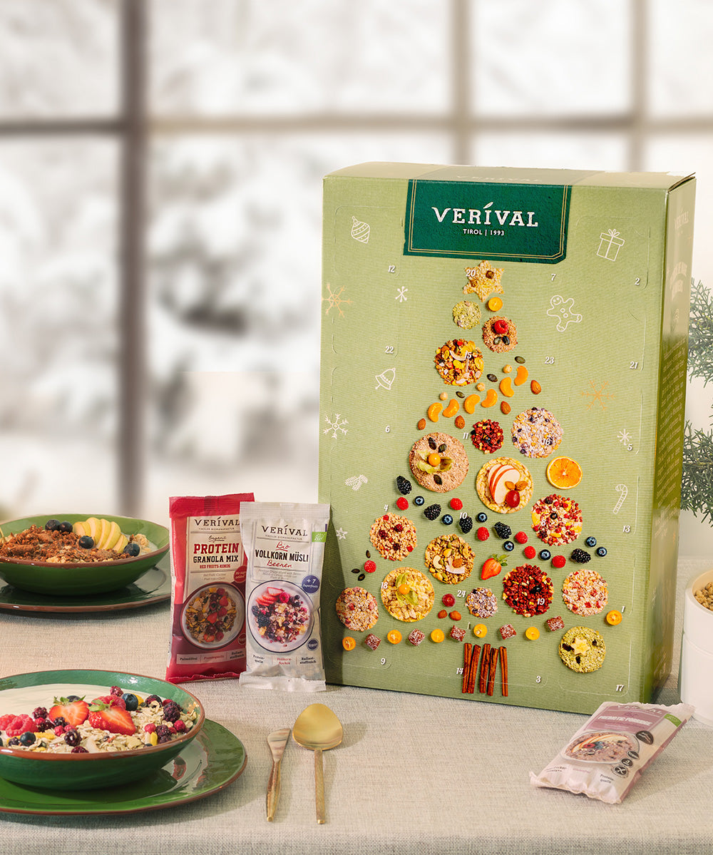 Eine Adventskalender-Box der Verival Bio GmbH, geschmückt mit getrockneten Früchten in Form eines Weihnachtsbaums, steht auf einem Tisch mit Schalen von Bio-Müsli und Vollkorngetreide, umrahmt von einem verschneiten Fenster im Hintergrund.