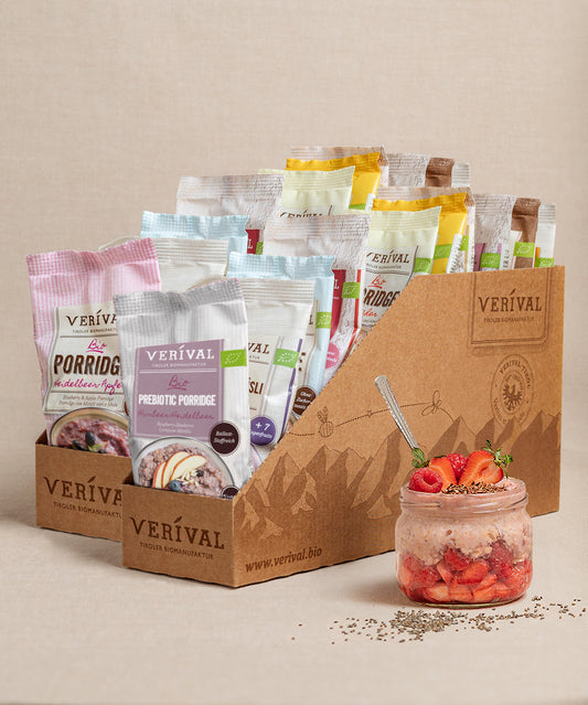 Die Große Frühstücks-Probierbox Top 25 der Verival Bio GmbH wird in Kartonbox präsentiert. Davor steht ein Glas mit Porridge, frischen Erdbeeren und Chiasamen, ideal für ein Bio-Frühstück.