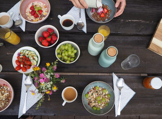 Ein Frühstückstisch mit Müslischüsseln, Erdbeeren, Weintrauben, geschnittenem Gemüse, Getränken, Tassen, Besteck, Servietten und einer kleinen Blumenvase. Eine Hand gießt Milch in eine Schüssel.