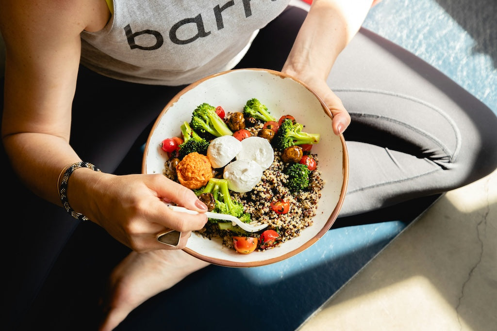 Eine Person in Trainingskleidung sitzt im Schneidersitz, hält eine Schüssel mit Quinoa, Brokkoli, Kirschtomaten und pochierten Eiern und isst mit einer Gabel.