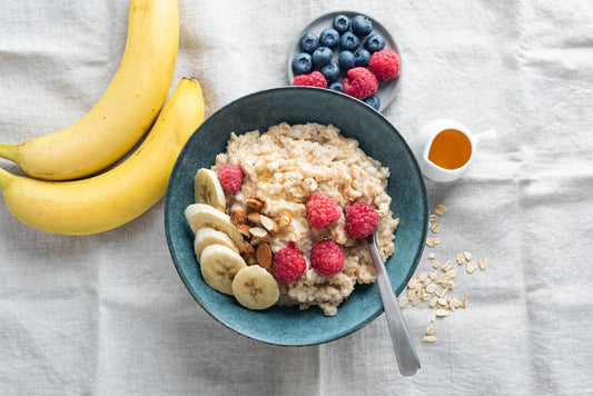 Eine Schüssel Haferbrei mit Bananenscheiben, Himbeeren und Nüssen, daneben zwei Bananen, ein Teller mit Blaubeeren und Himbeeren, ein kleines Kännchen Honig und verstreute Haferflocken auf einem Tuch.