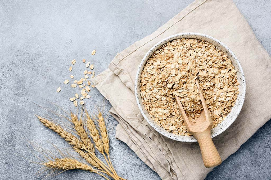 Eine Schüssel mit Haferflocken und einem Holzlöffel steht auf einem beigen Tuch neben Weizenhalmen und verstreutem Hafer auf einer grauen Oberfläche.
