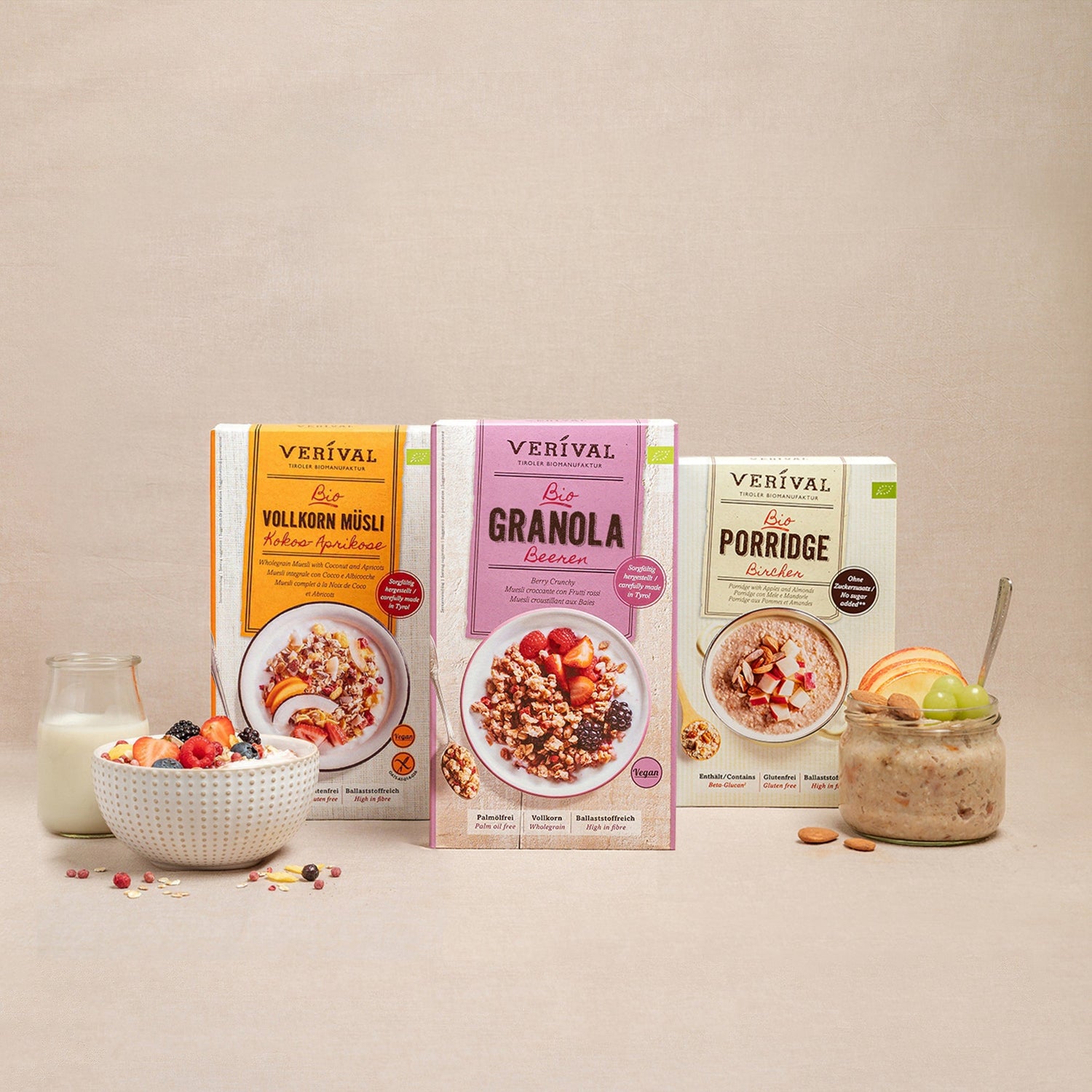 Three muesli boxes from Verival: Whole Grain Muesli, Granola Berries, and Porridge. Pictured with a bowl of muesli, a bowl of porridge, a glass of milk, and fruit on a beige background.