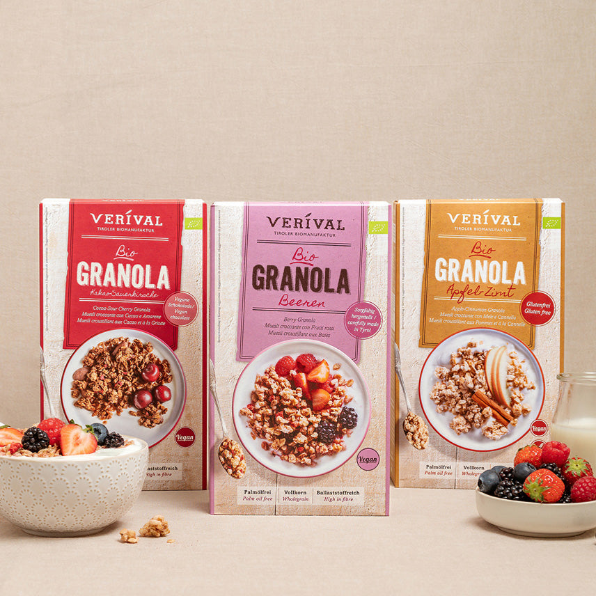 Three boxes of Verival granola in various flavors stand upright on a beige surface, with a bowl of granola and mixed berries in front of them.