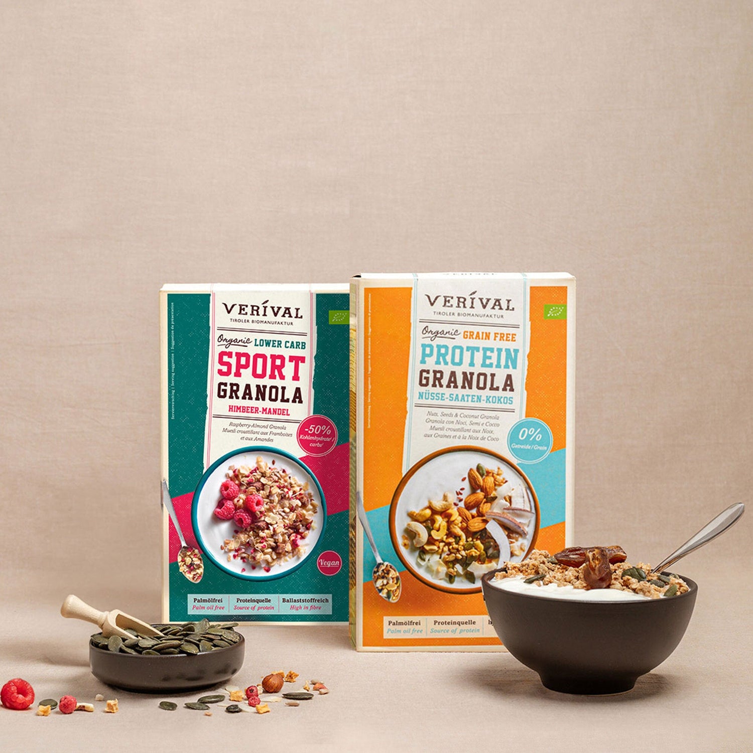 Two boxes of Verival granola (Sport Granola and Protein Granola) stand behind a bowl of granola with a spoon and a small bowl of seeds against a neutral background.