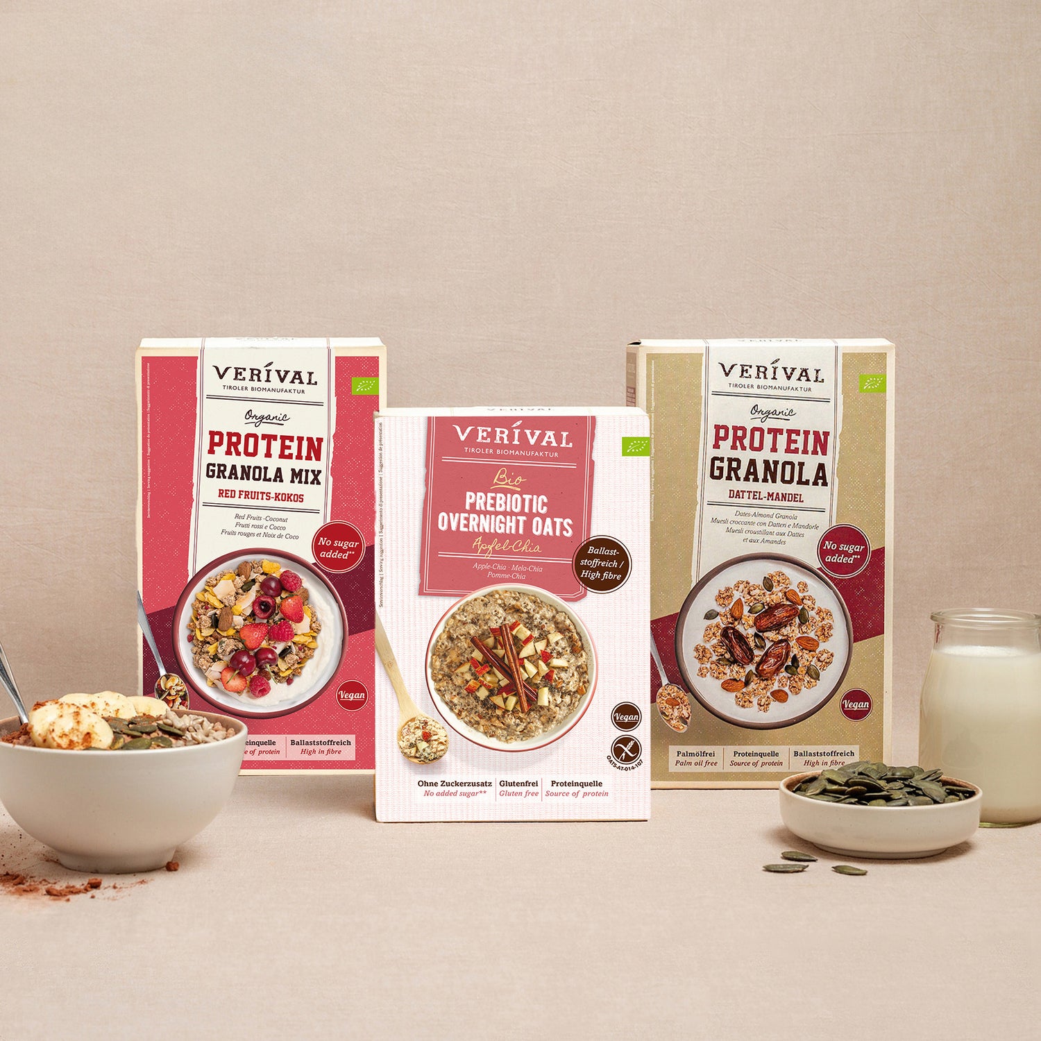 Drei Packungen Verival Frühstückscerealien, darunter Protein Granola Mix, Prebiotic Overnight Oats und Protein Granola. Vorne stehen eine Schüssel Müsli, ein Löffel, Nüsse und ein Glas Milch.