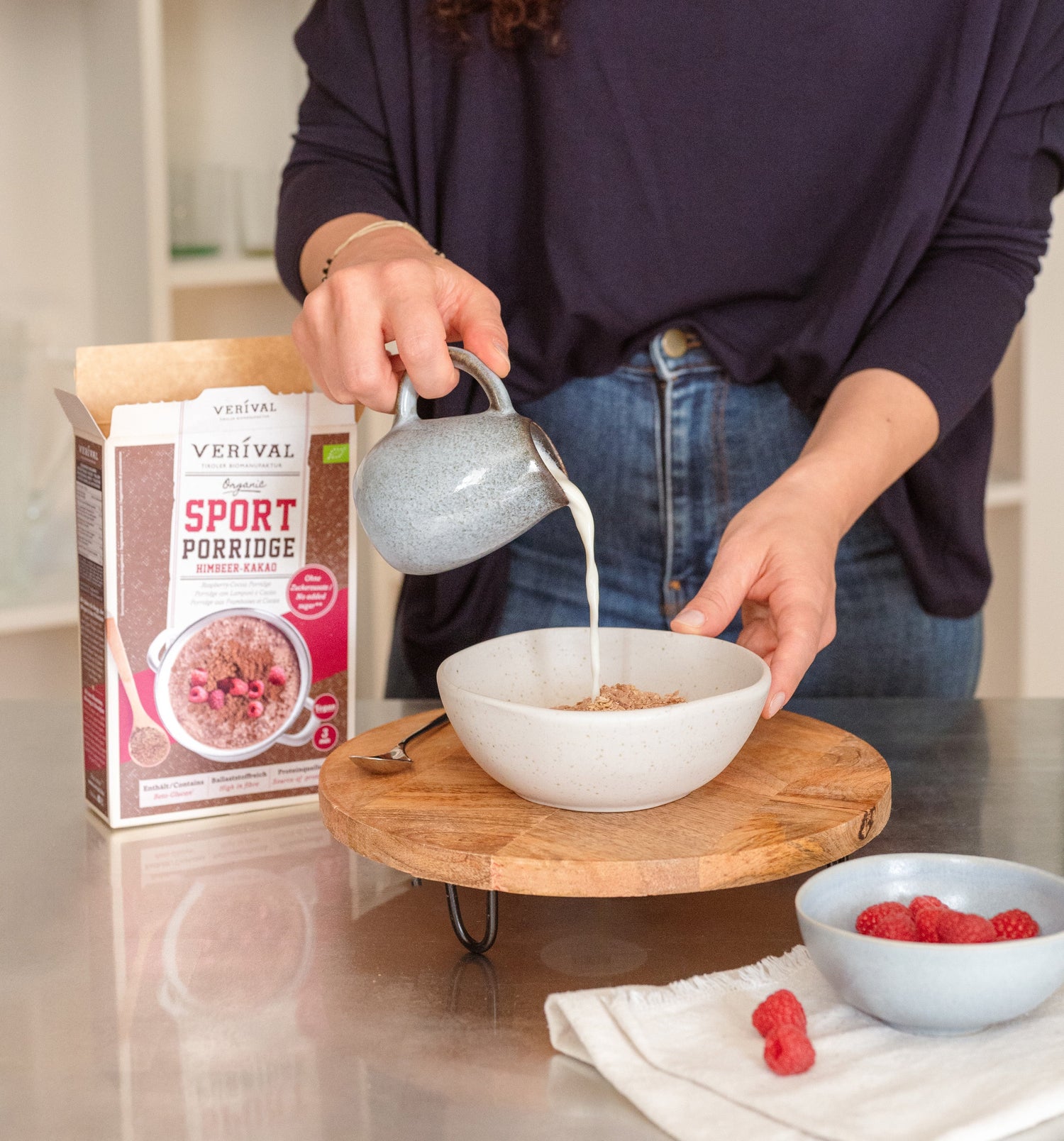 A person pours milk into a bowl of porridge next to a box labeled Sport Porridge and a small bowl of raspberries on a wooden tray.