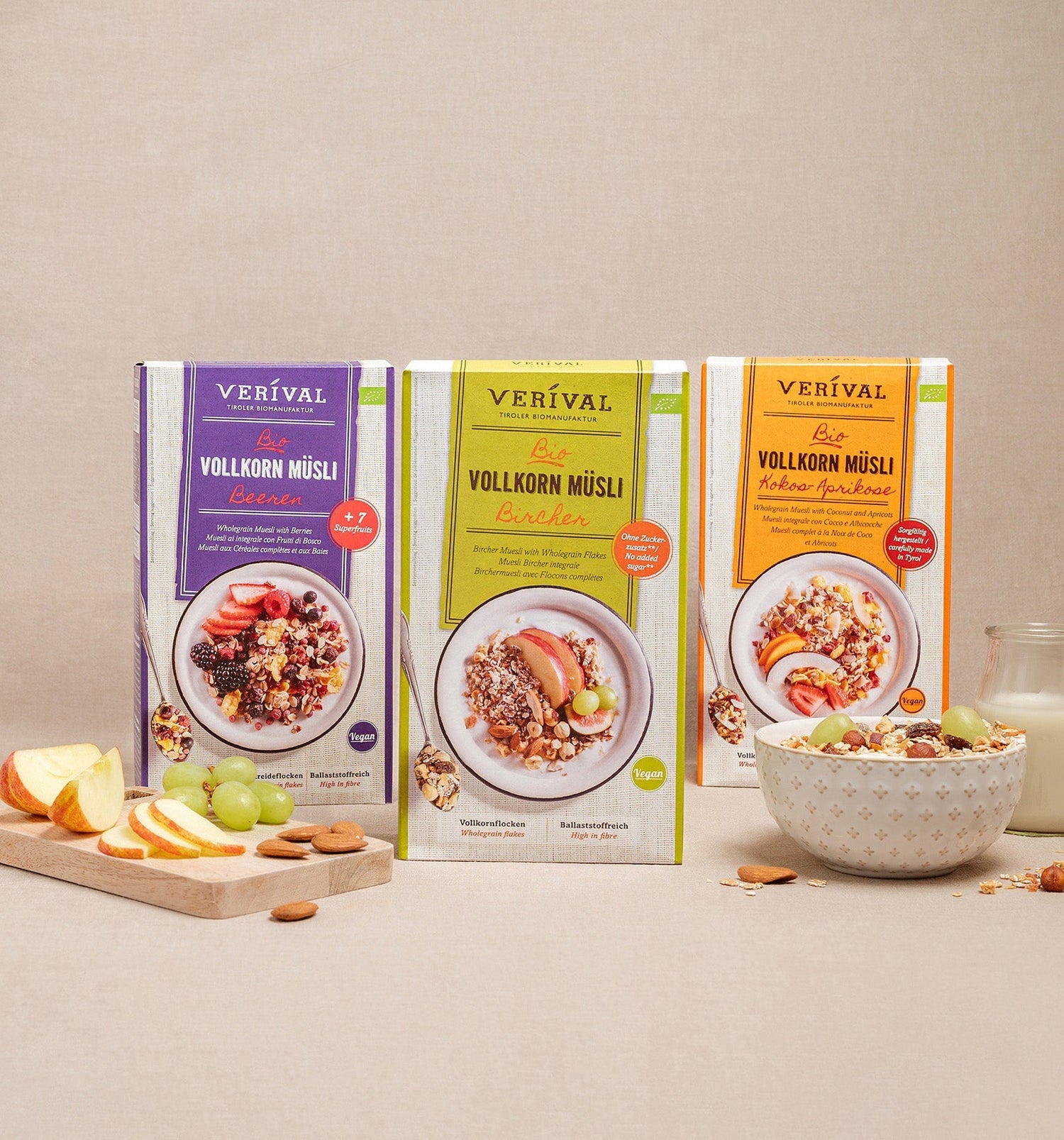 Three boxes of Verival organic wholegrain muesli stand upright, in front of them a bowl of muesli, apple slices, grapes, a spoon and a glass of milk.