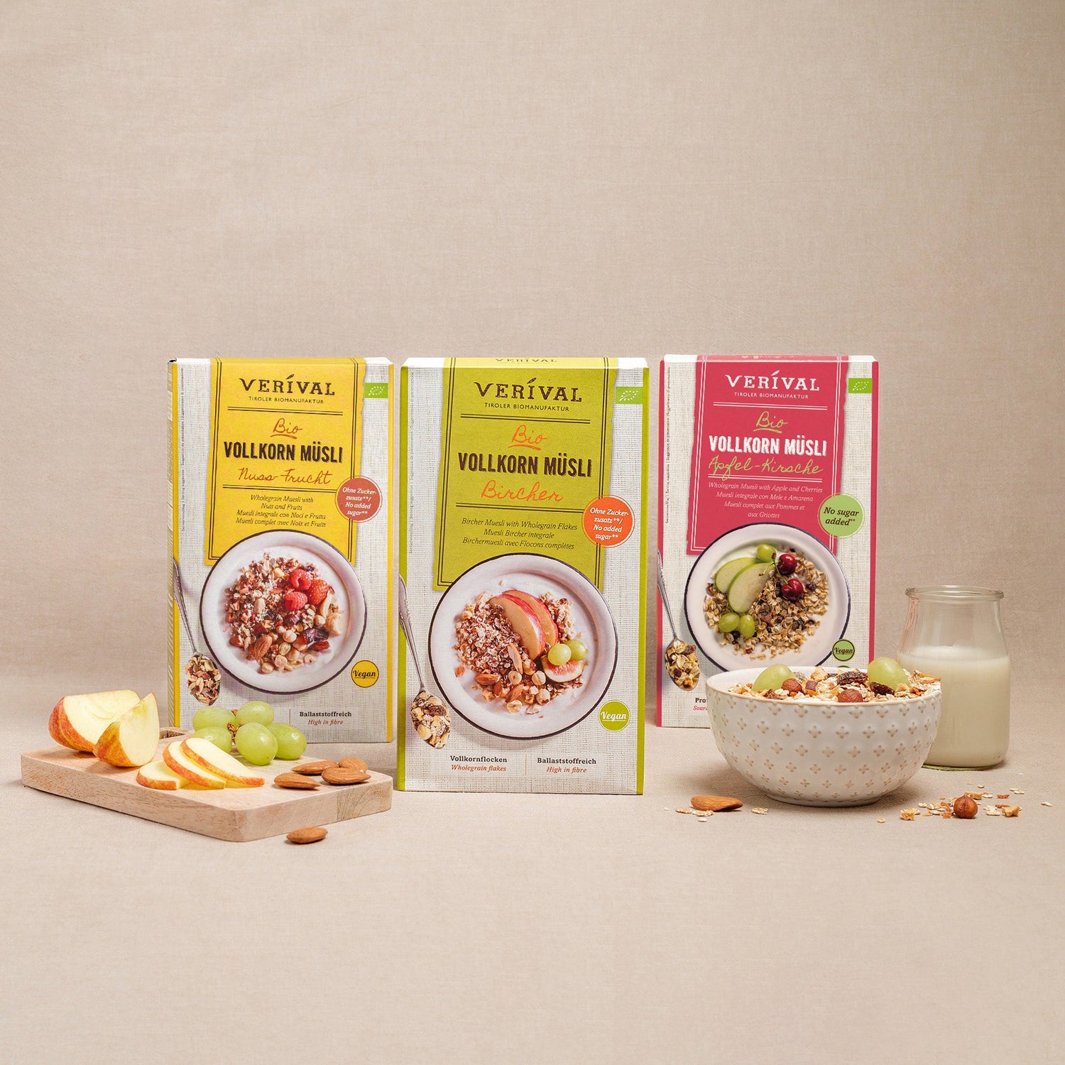 Three boxes of Verival wholegrain muesli in various flavors are presented upright with a bowl of muesli, a glass of milk, apple slices, and grapes in front of it.