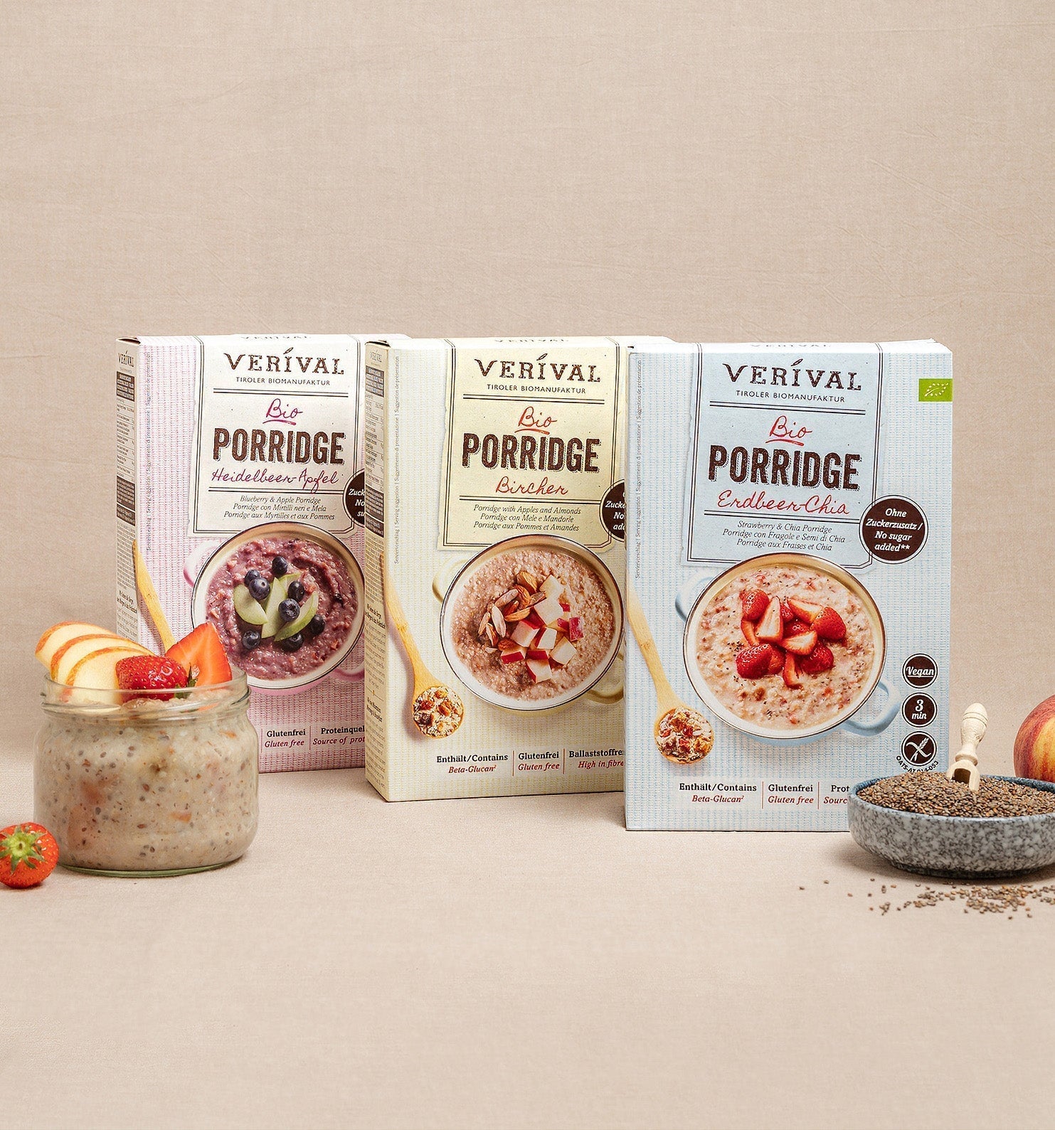 Three boxes of Verival Organic Porridge (apple, Bircher, and chia varieties) are presented with a glass of porridge, fresh fruit, chia seeds, and an apple on a beige background.