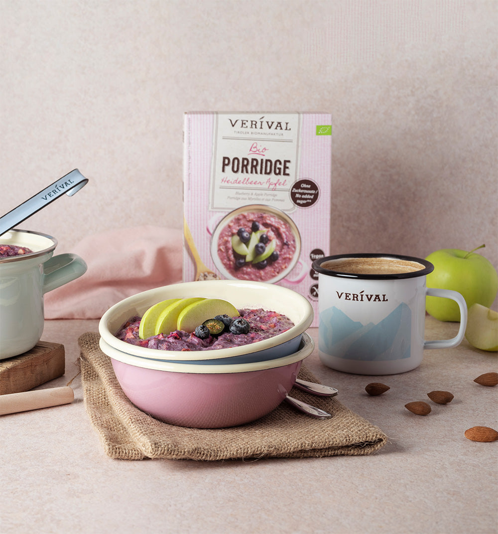 Three boxes of Verival organic porridge in different flavors stand next to a jar of ready-made porridge, strawberries, apple slices and a bowl of chia seeds.