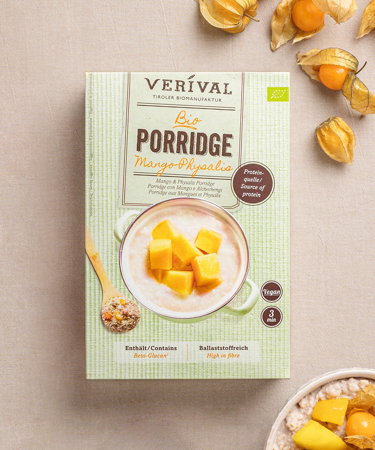 Eine Packung Mango-Physalis Porridge von Verival Bio steht auf einem beigen Untergrund. Die Schüssel ist übervoll mit beta-glucanreichem Porridge, garniert mit frischer Mango und verstreuten, leuchtenden Physalis-Körnern. 