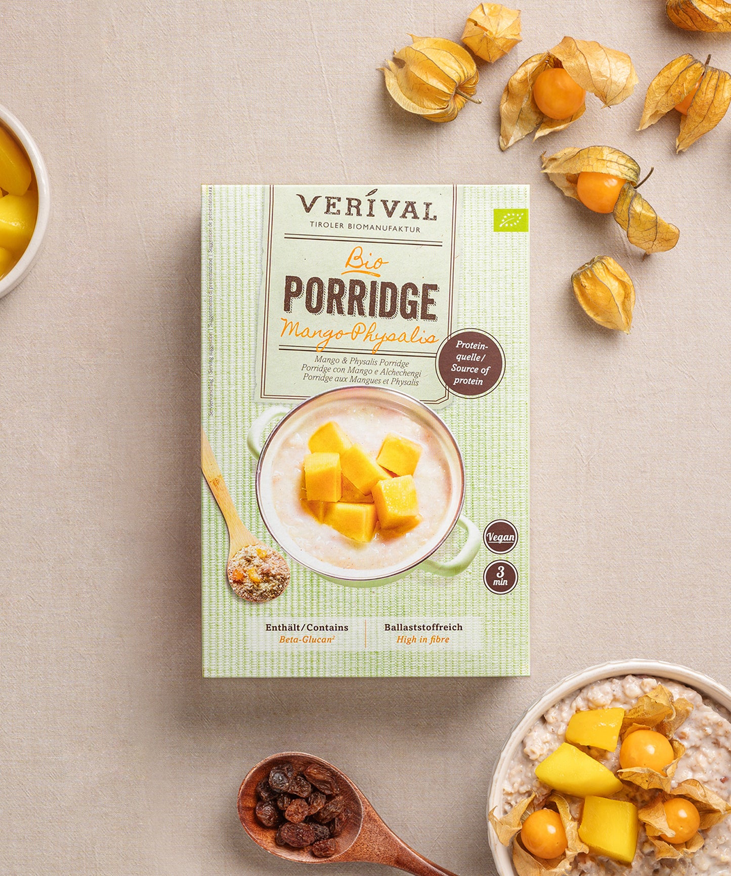 Eine Paket Mango-Physalis Porridge von Verival Bio steht auf einer beigen Fläche, umgeben von Physalisfrüchten, einer Schüssel an Beta-Glucan-reichem Porridge und einem kleinen Teller Rosinen.
