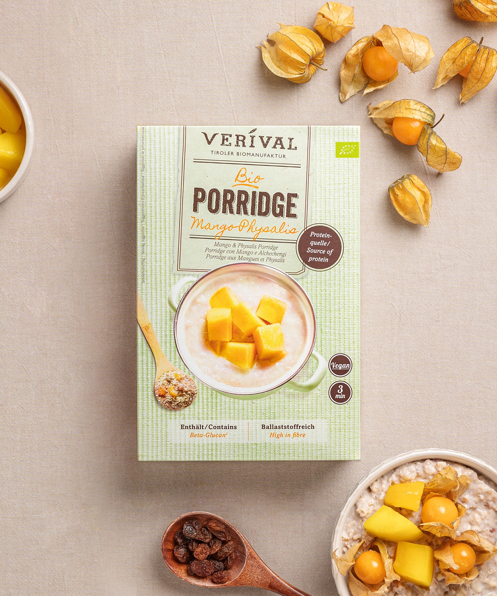 Eine Paket Mango-Physalis Porridge von Verival Bio steht auf einer beigen Fläche, umgeben von Physalisfrüchten, einer Schüssel an Beta-Glucan-reichem Porridge und einem kleinen Teller Rosinen.