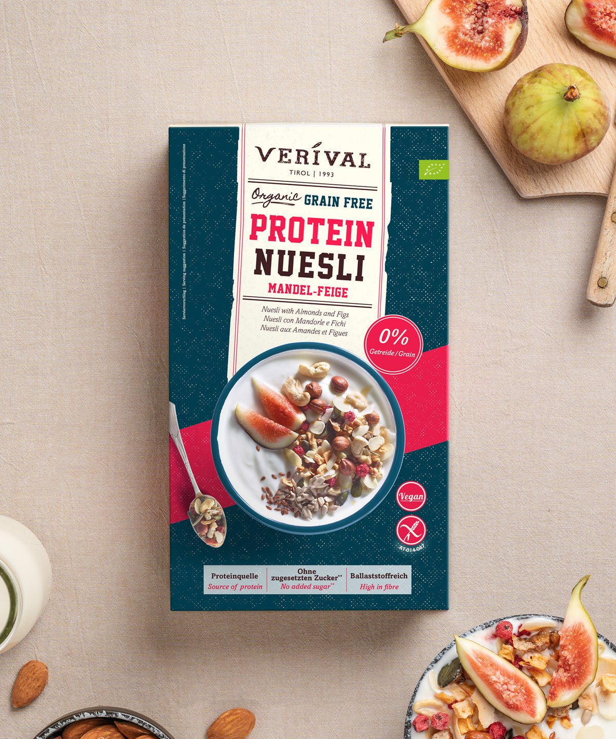 Eine Packung Verival Bio Bio Mandel-Feige Grain Free Protein Nüsli, umgeben von frischen Feigen, Nüssen und Schalen mit Müsli.