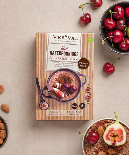 Eine Packung Sauerkirsche-Kakao-Haferflockenporridge von Verival Bio liegt neben Kirschen und Mandeln auf einem Tisch. Auf der Verpackung steht „Ohne Zuckerzusatz“ und eine Zubereitungszeit von 3 Minuten. 