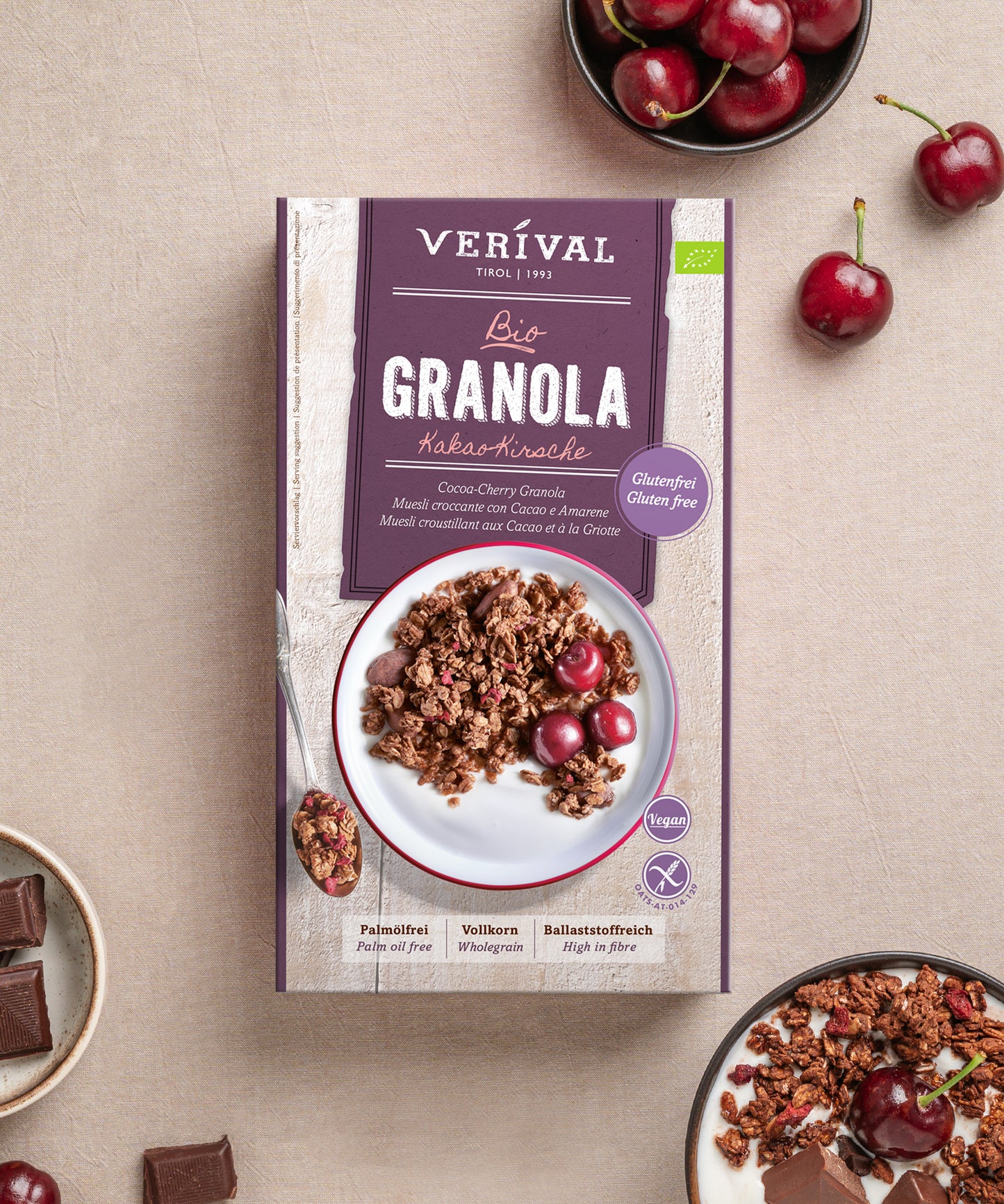Eine Schachtel Bio Kakao-Kirsche Granola glutenfrei der Verival Bio GmbH ist umgeben von frischen Kirschen, Schokoladenstückchen und einer Schale Granola auf einer hellen Fläche abgebildet.
