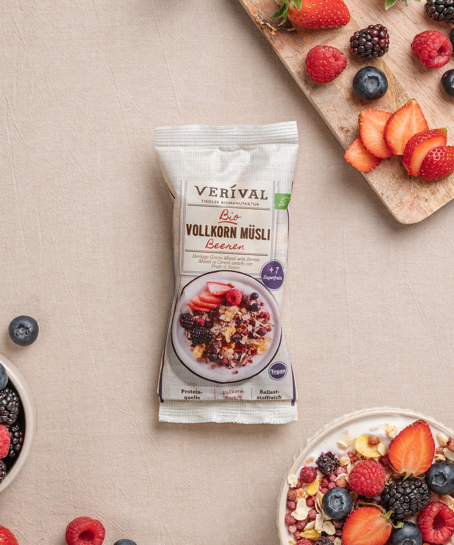 Ein Einzelportionspackung Beeren Vollkorn Müsli von Verival Bio, umgeben von Schalen mit diesem ballaststoffreichen Müsli und einem Holzbrett mit gemischten Beeren. 