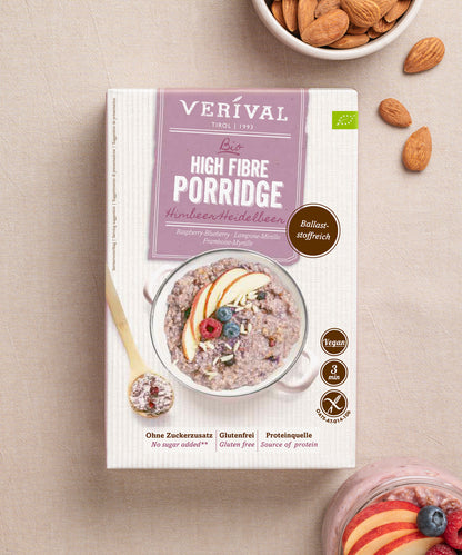 Der Bio Prebiotic Porridge Himbeer-Heidelbeer der Verival Bio GmbH auf einer leichten Unterlage, umgeben von Mandeln und einer Schale ballaststoffreichem, veganem Porridge, garniert mit frischen Obstscheiben - das ideale gesunde Frühstück.