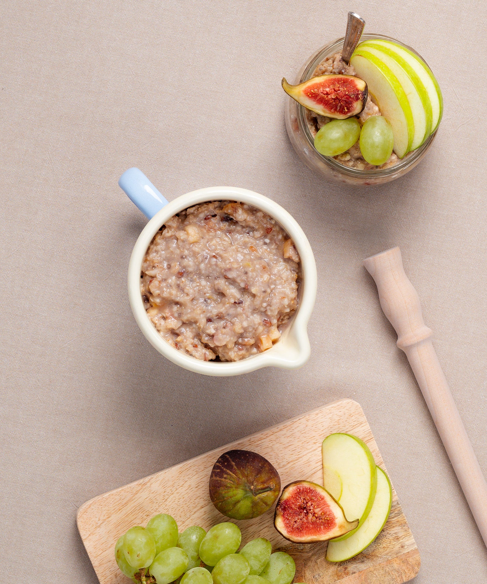 Auf einer beigen Fläche steht steht der Riess Schnabeltopf. Auf einem Holzbrett liegen Apfelscheiben, Trauben und Feigen neben dem Porridge-Rührstab. 